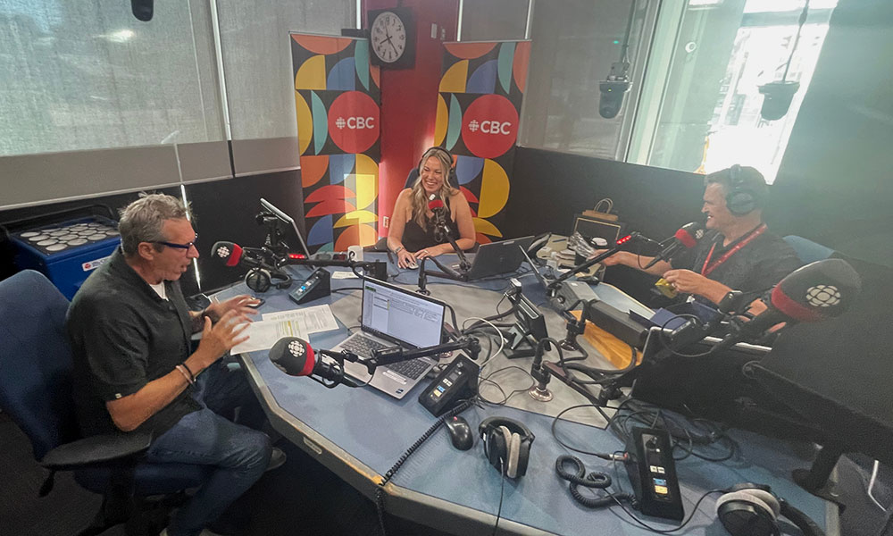 Three individuals seated around a table in a CBC radio studio, speaking into microphones with broadcasting equipment and CBC banners in the background.