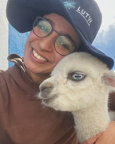 a woman wearing a blue bucket hat and glasses smiles while she hugs a white baby alpaca