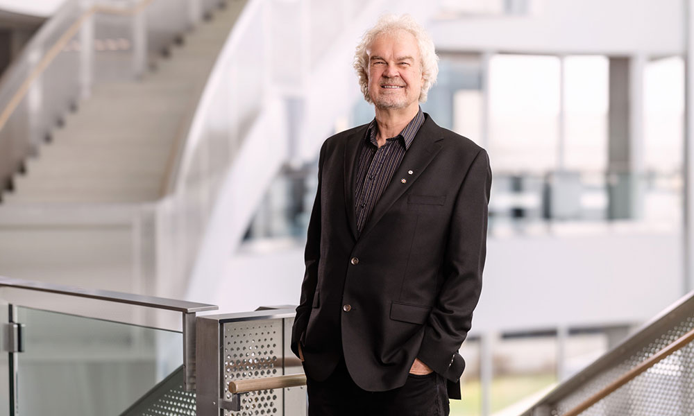 an older man in a black blazer and dark striped open-collared short poses for a photo in a modern-looking building