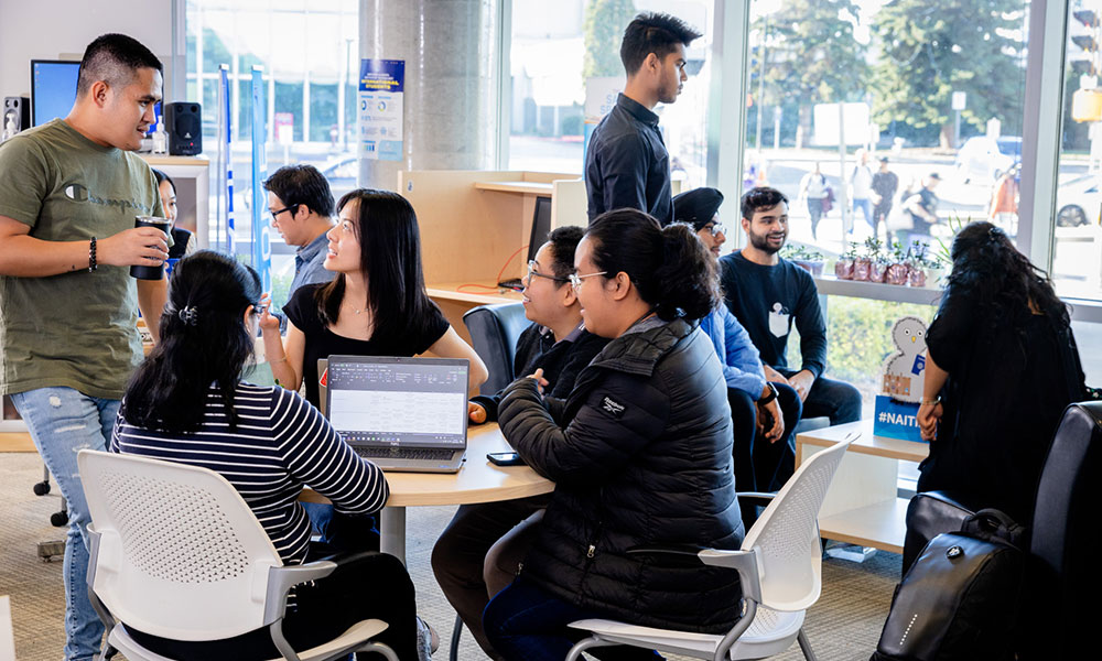 Group of people gathered around a table with laptops in a bright room near large windows.