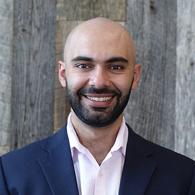 A person wearing a dark blazer over a light shirt stands in front of a wooden textured background, looking directly at the camera.