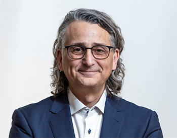 man wearing a blue blazer and white shirt buttoned to the collar without a tie. he has glass and wavy, shoulder length grey hair