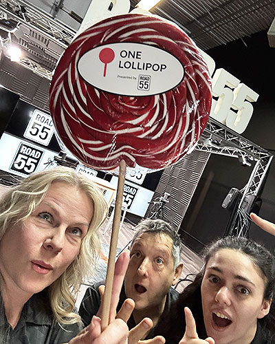 Several people pose behind a large red‑and‑white swirled lollipop labeled “ONE LOLLIPOP” in a studio setting with multiple “ROAD 55” signs in the background.