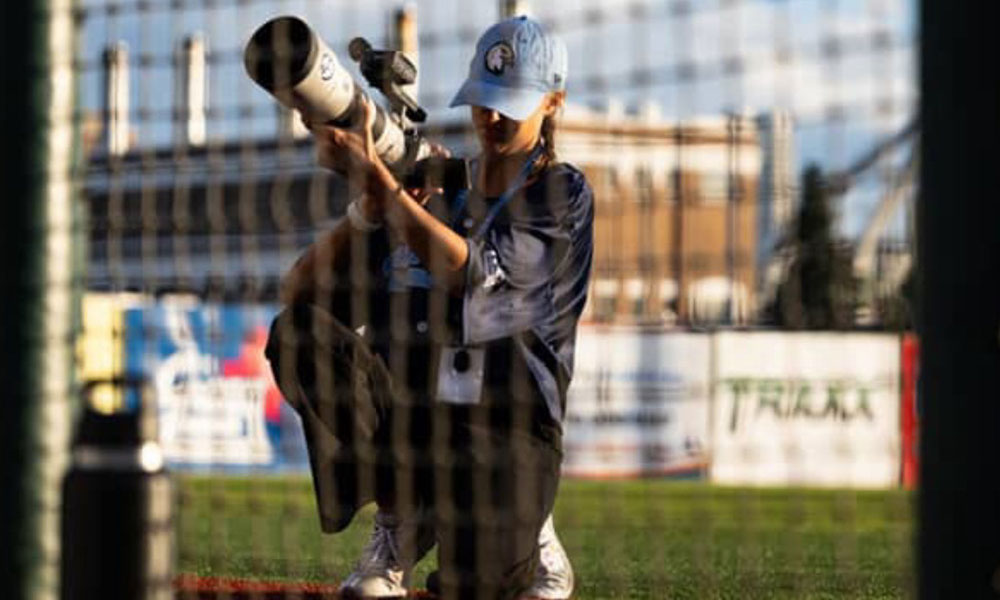 Person kneeling on a sports field behind a net, holding a large telephoto camera lens aimed forward, wearing a light blue cap and dark shirt with a lanyard.