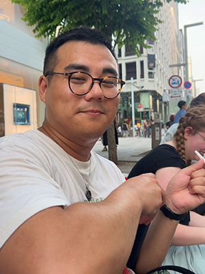 Person wearing glasses and a white t-shirt sitting outdoors in an urban area, pointing off-camera while holding an object. Shops and people are visible in the background, with another person partially seen beside them.