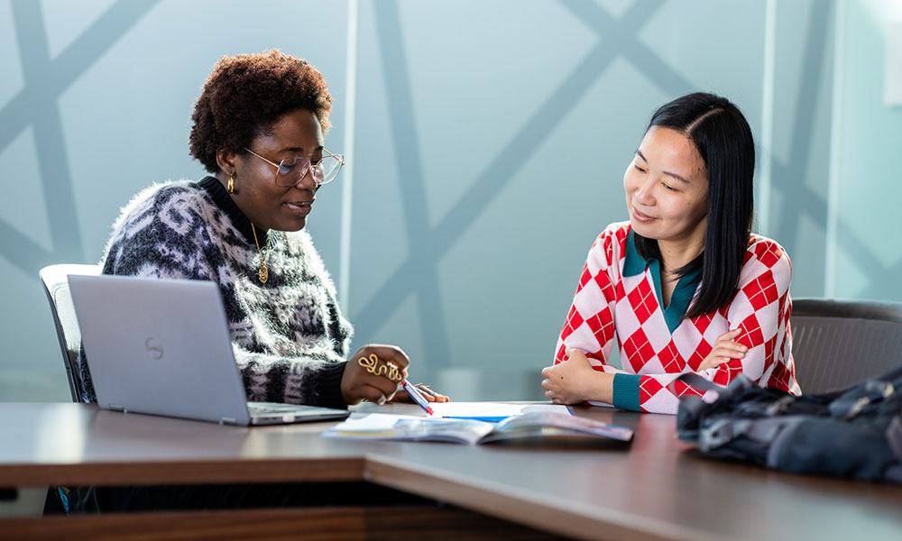 Two people sitting at a table with a laptop and open documents, discussing paperwork in a bright indoor setting.