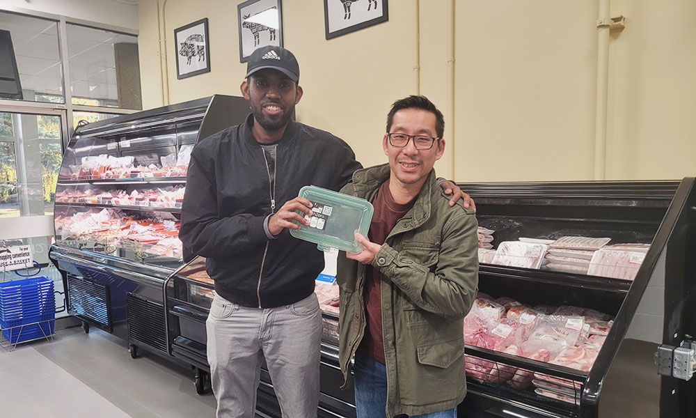 Two people standing in front of a refrigerated meat display in a grocery store, holding a green reusable DishZero container together. The background shows multiple shelves stocked with packaged meat, a row of blue shopping baskets on the left, and framed diagrams of meat cuts on the wall above.