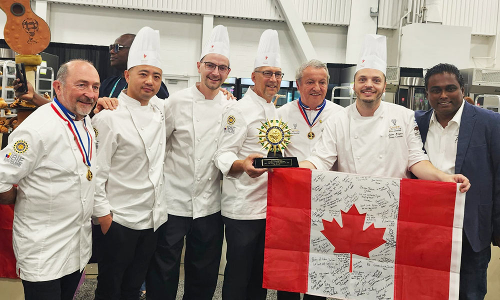 Group of chefs in white uniforms and tall hats standing together, holding a trophy and a signed Canadian flag at an indoor event.