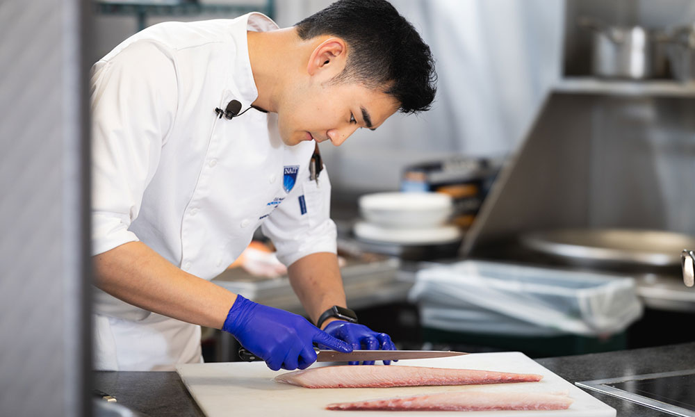 Person in a professional kitchen slicing raw fish fillets on a cutting board while wearing blue gloves and a white chef’s jacket.