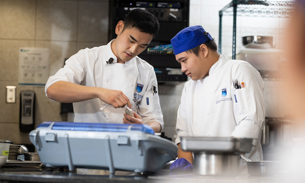 Two people in white chef jackets stand in a commercial kitchen, closely examining an item on a counter beside food-service equipment.