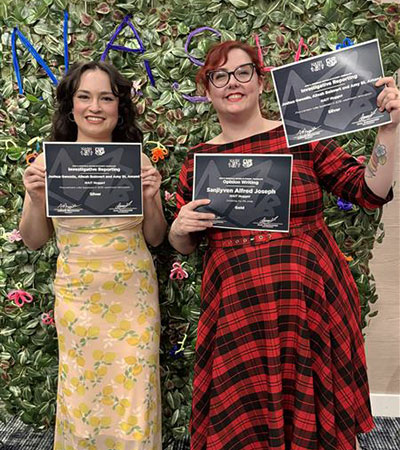 Two people stand in front of a leafy wall display holding framed certificates, with colorful lettering visible in the background.