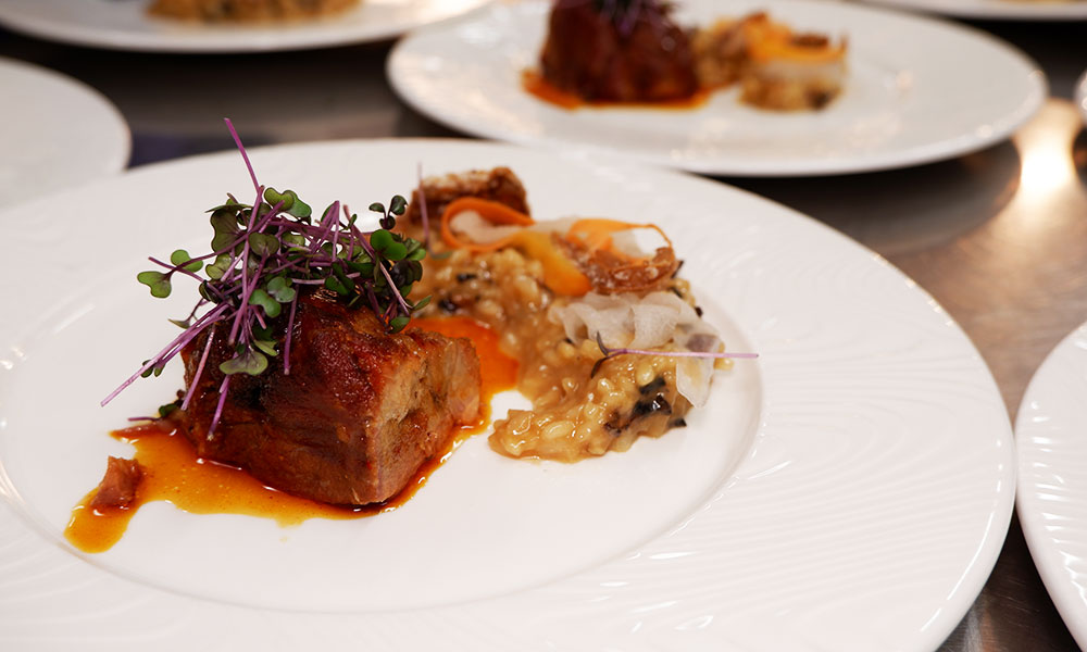 A plated dish featuring a piece of braised meat topped with microgreens, served alongside a creamy risotto with mushrooms and thin vegetable slices. The plate is white, and other similar plates are visible in the background on a stainless steel surface.