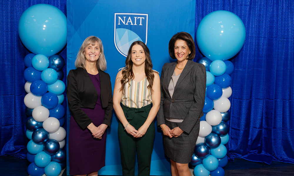 three women pose for a photo. they are wearing business and formal attire. behind them is a blue background and blue balloons.