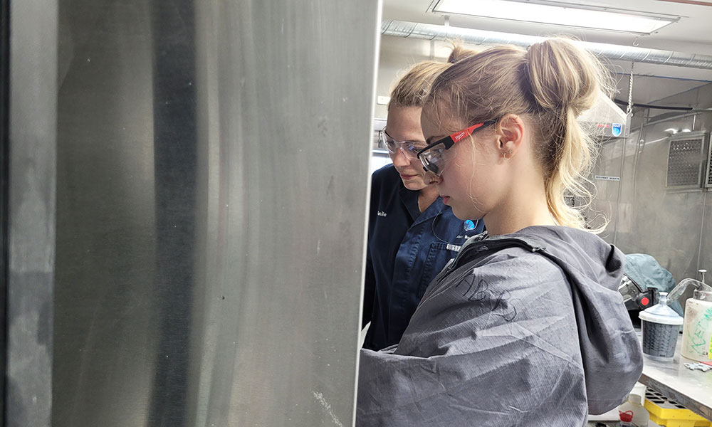 Two individuals wearing lab coats and safety glasses working in a laboratory setting. One person in the foreground has their hair tied up and is focused on a task out of frame. The background includes lab equipment and containers.
