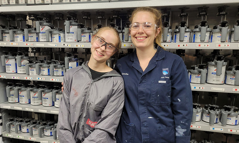 Two individuals wearing protective eyewear and work attire standing in front of shelves filled with labeled containers. One wears a gray jacket with a red logo, and the other wears a dark blue jacket with name tags reading "Cecile" and "Axalta Body." The setting appears to be an industrial workspace involving paint.