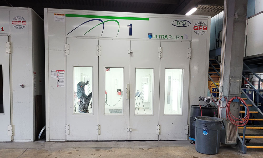 Industrial paint booth labeled "ULTRA PLUS 1" with four glass windows showing a person in protective coveralls inside. Warning signs are posted on the left, and a trash bin with coiled hoses is on the right. The booth is part of a larger industrial facility with nearby stairs and machinery.