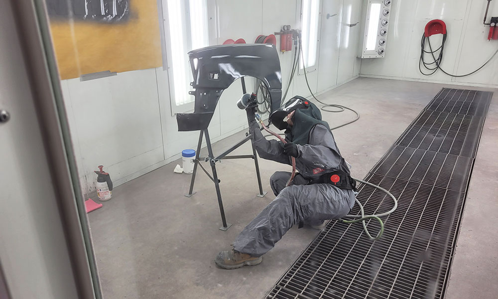 Person in full protective gear spray painting a car fender inside a well-lit industrial paint booth. The fender is mounted on a stand, and the person is using a spray gun connected to an air hose.