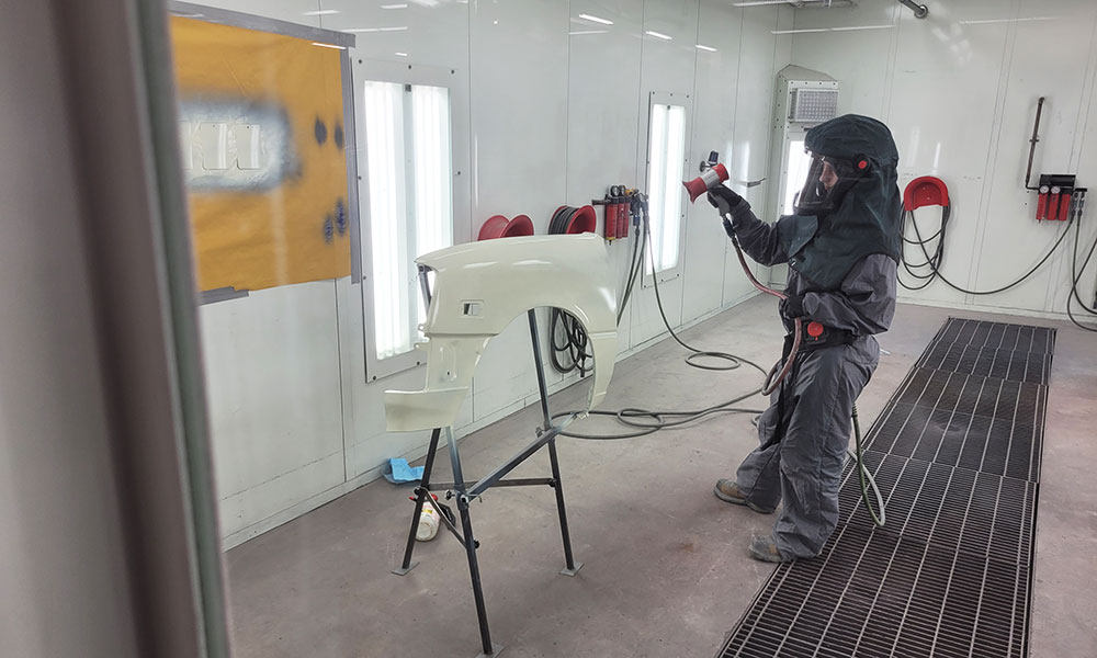 Person in full protective gear spray painting a white car fender mounted on a stand inside a professional paint booth. The booth is brightly lit with visible ventilation systems.