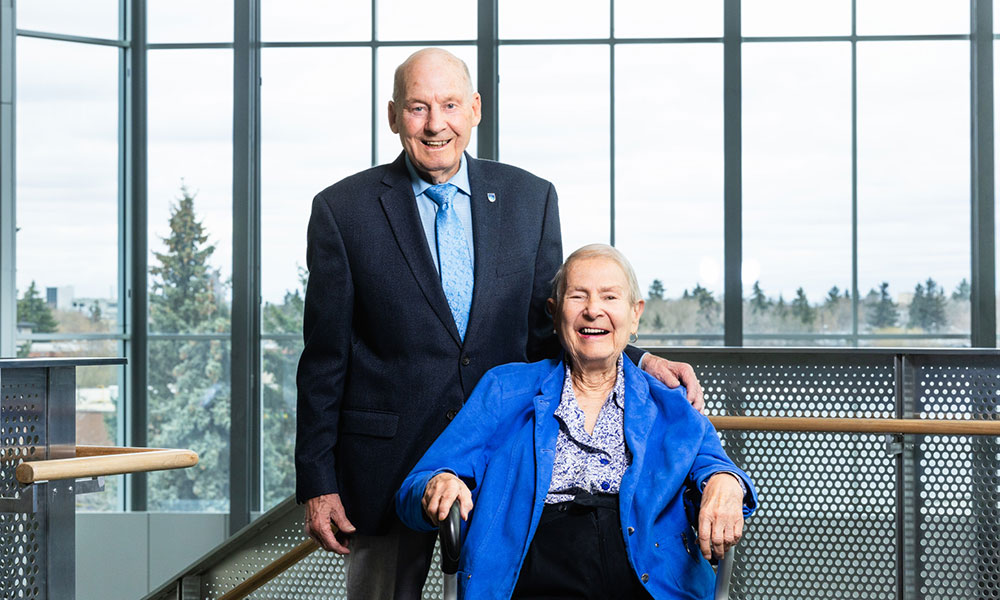 Two people posed indoors on a modern staircase landing, one standing in a suit and tie, the other seated in a wheelchair wearing a bright blue jacket. Large windows behind them show trees and an overcast sky.