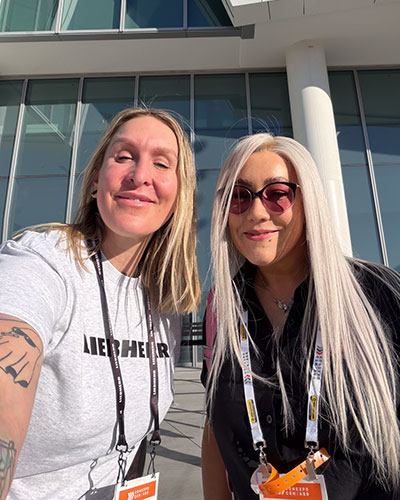 Two women wearing conference lanyards smile for a selfie outside a modern glass building in bright sunlight.