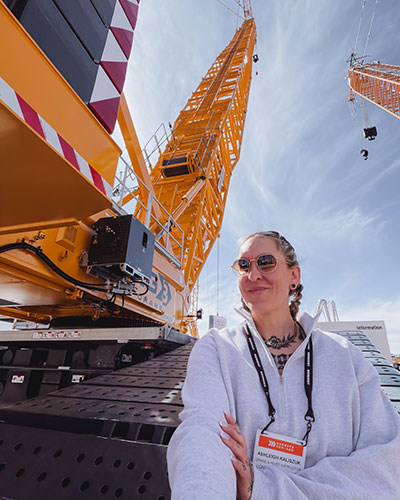 Person wearing a lanyard stands confidently in front of a large yellow construction crane at an industrial site under a blue sky.