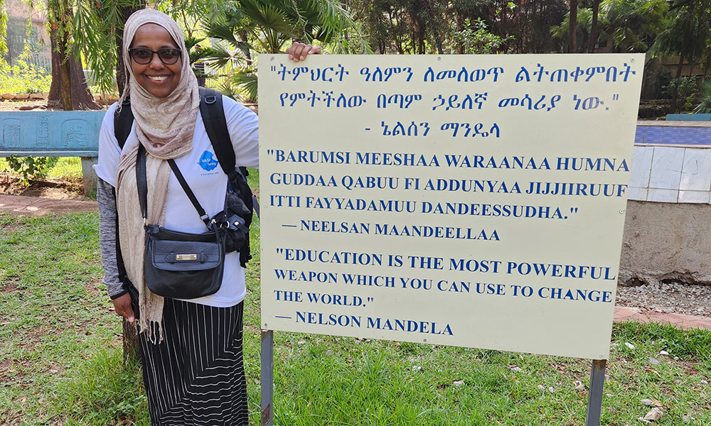 Person wearing glasses and a head covering stands outdoors beside a sign displaying a Nelson Mandela quote about education in multiple languages.