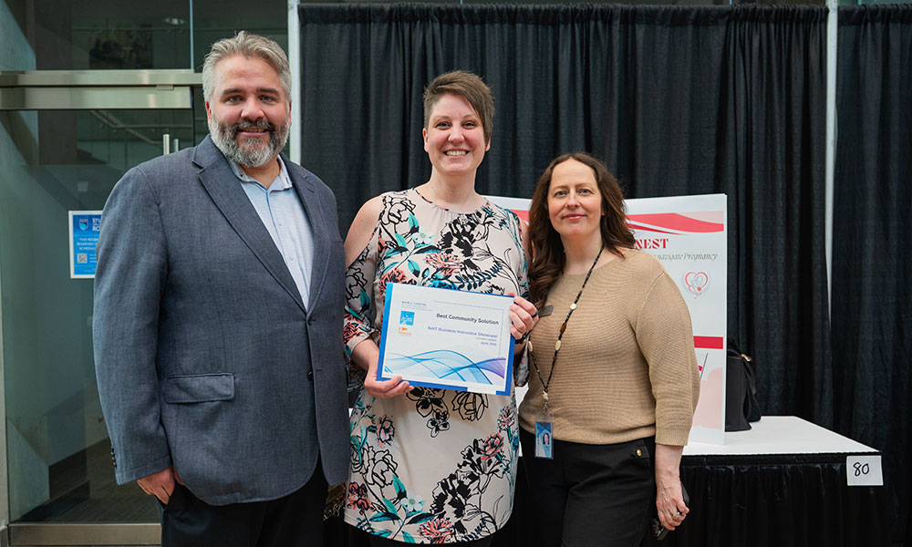 a photo of a man in a blue blazer, a woman in a white dress with a flower pattern, and a woman in a brown sweater. the woman in the dress, in the middle, holds a certificate.