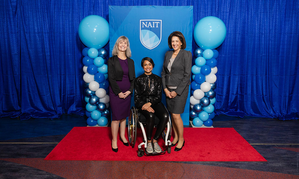 three women pose for a photo. they are wearing business and formal attire. behind them is a blue background and blue balloons. the woman in the middle sits in a wheelchair