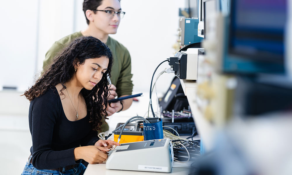 Two individuals in a laboratory working with electronic equipment, including an oscilloscope and a tablet, surrounded by cables and devices in a technical learning environment.