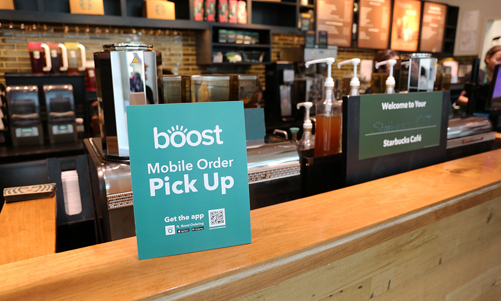 Counter at a Starbucks café featuring a sign promoting mobile order pickup and app downloads, with another sign in the background reading 'Welcome to Your Starbucks Café'.
