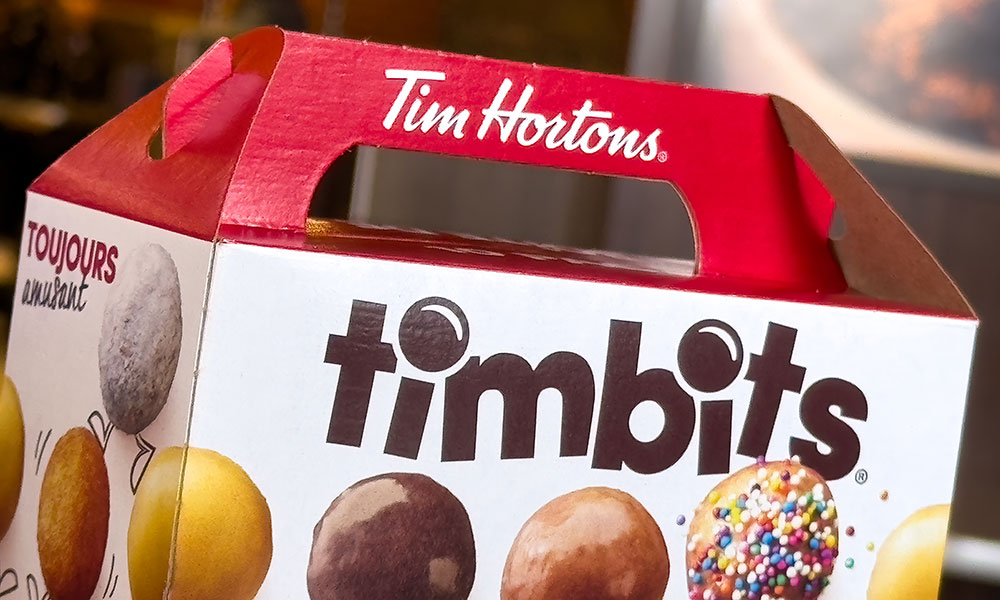 A box of Timbits from Tim Hortons with a red handle and 'Tim Hortons' written in white. The front displays 'timbits' in bold black letters, surrounded by images of doughnut holes, and includes French text 'TOUJOURS frais' on the side.
