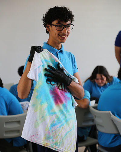 A person wearing a blue polo shirt and black gloves holds up a colorful tie-dyed t-shirt with the partially visible word 'EQUALITY.' In the background, other people are seated at tables participating in activities.
