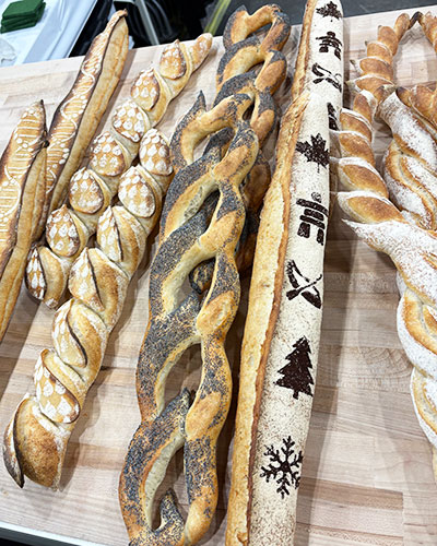 Assorted artisan baguettes displayed on a wooden surface, featuring intricate designs such as leaf-shaped cuts, twisted shapes, poppy seed coatings, and stenciled patterns of trees and snowflakes on the crust.