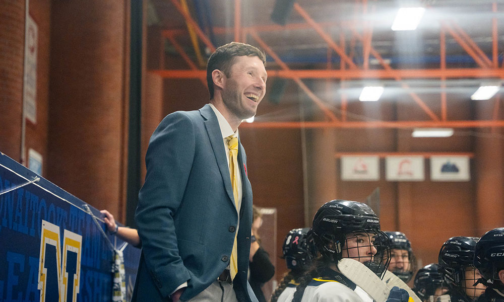 A person wearing a blue suit jacket and yellow tie stands behind the bench during a hockey game, with several players in black helmets seated in front. The background shows an indoor arena with orange beams and bright overhead lights.