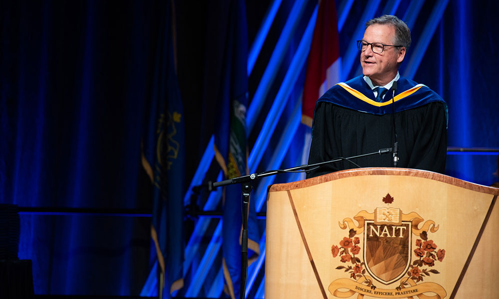 Person in academic regalia standing at a podium with the NAIT emblem, during a graduation or academic ceremony. Background includes blue lighting and several flags.