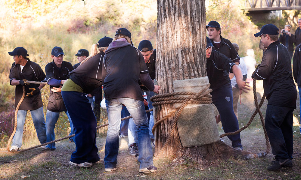 A group of people dressed in dark clothing and caps work together outdoors, pulling a rope wrapped around a large padded tree with multiple ropes tied to it, suggesting a teamwork or physical challenge activity.
