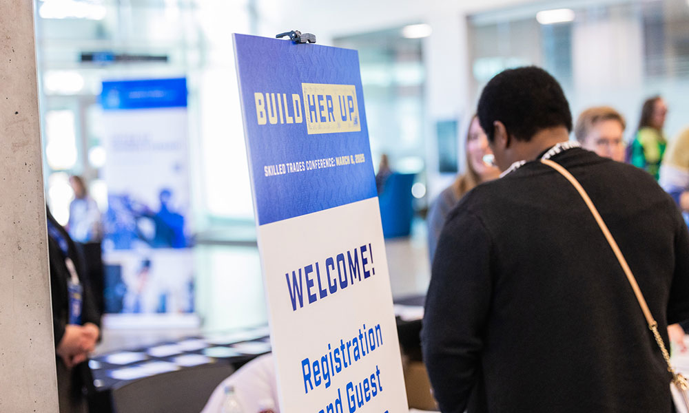 A large “BUILD HER UP” conference welcome sign stands at a registration area while attendees gather nearby.