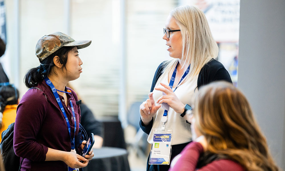 Two people wearing event lanyards talk together in a conference setting while another person sits in the foreground.