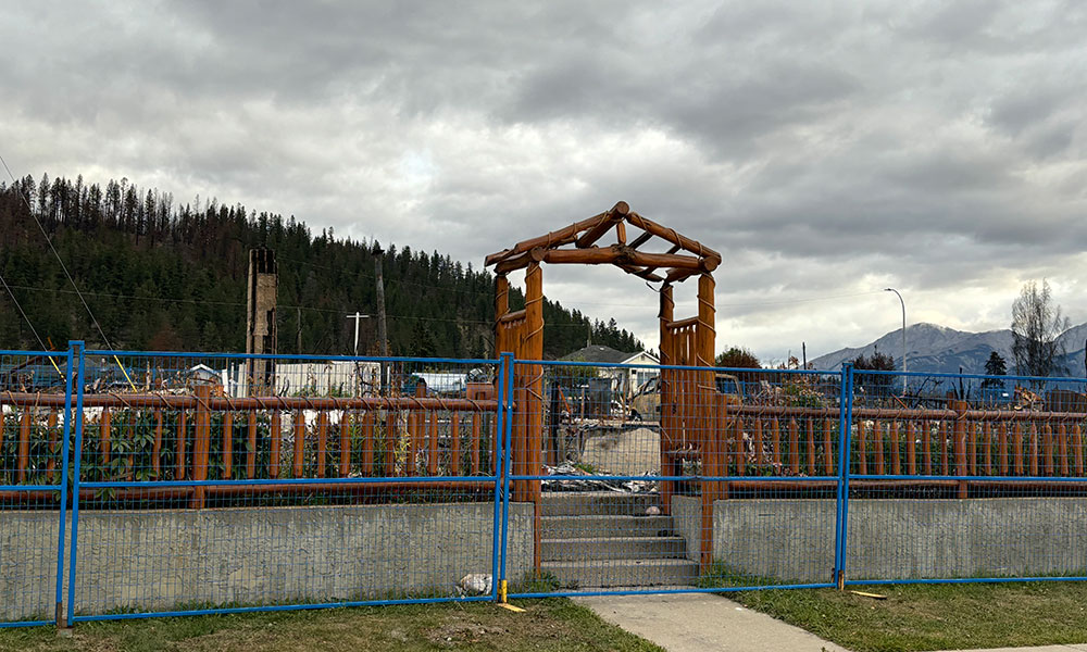 A wooden garden arbor stands intact amidst the ruins of a burned-down house. Blue fencing surrounds the property, with a concrete base and steps leading up to the arbor. The background shows more burnt structures and a forest on a hill under a cloudy sky.