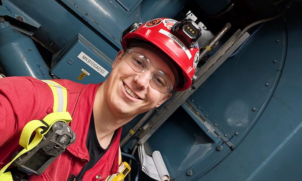 Person in red helmet with stickers and headlamp, wearing red uniform and yellow safety harness, standing in front of industrial equipment.