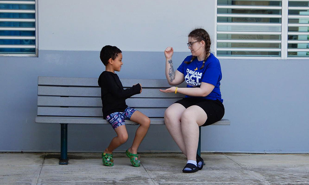 An adult and a child sitting on a bench outside a building. The adult wears a blue shirt with 'DREAM ACT ACHIEVE' and black shorts, while the child wears a black long-sleeve shirt, patterned shorts, and green sandals. They appear to be engaged in conversation or an activity. The background features windows with horizontal blinds and a grey wall.