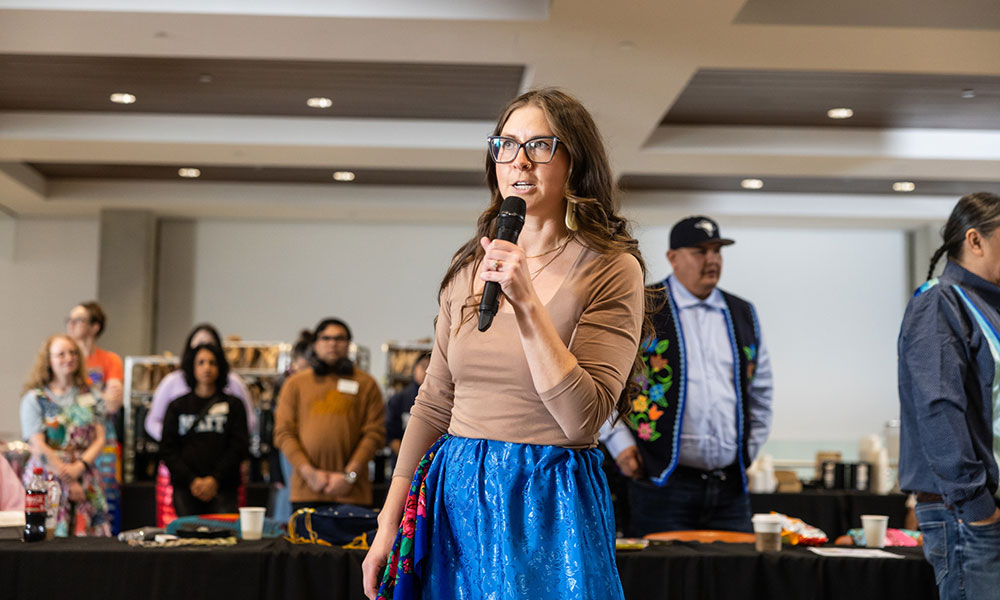 A person holding a microphone speaks to a gathered group at an indoor round dance event.
