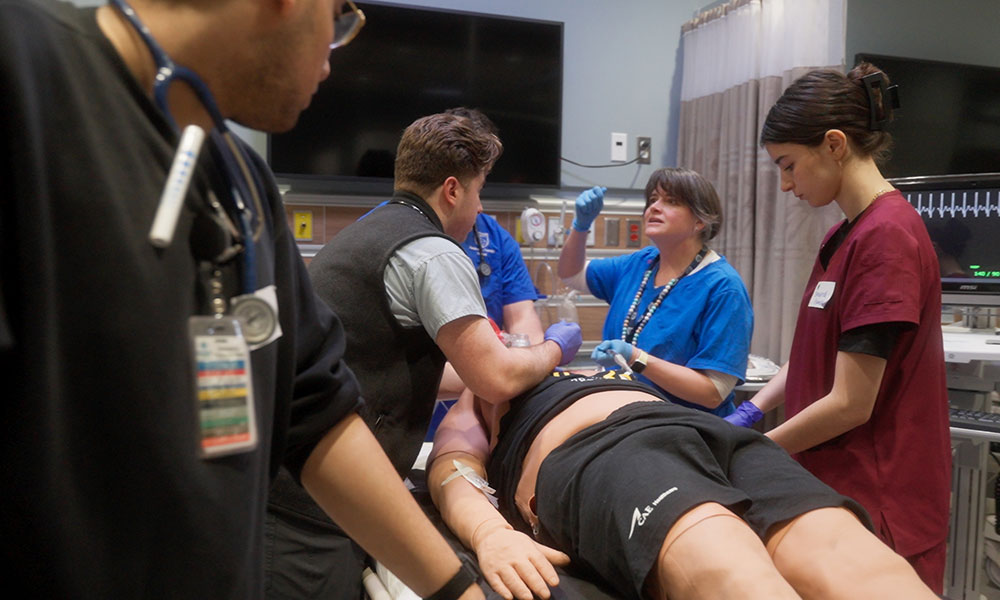 health care students around a patient in a bed in a simulated hospital room