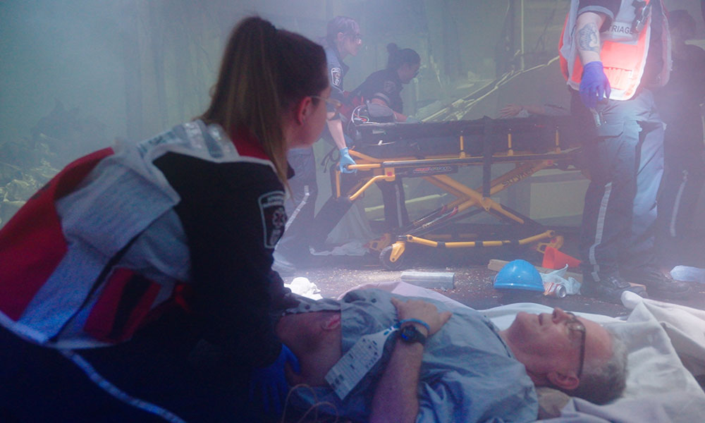 a paramedic student kneels by an actor patient and looks through a smokey scene as colleagues work around them