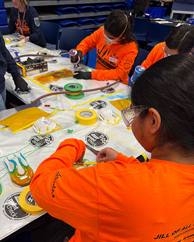 Participants in bright orange “Jill of All Trades” shirts work at a table covered with protective sheets, tape, and paint supplies. One person in the foreground, wearing safety goggles, applies tape to a small car panel while others paint in the background.