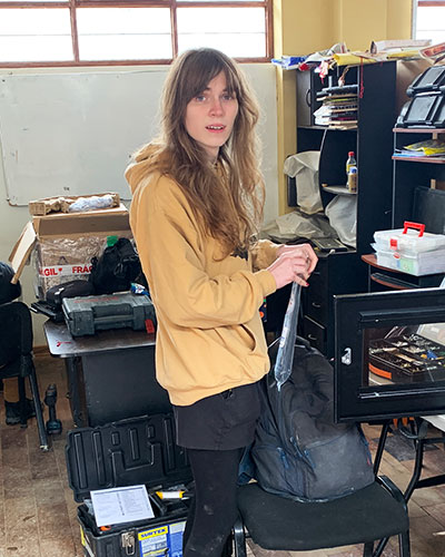 a woman in a yellow hoodie holds a small plastic bag in an office full of papers, equipment and tools