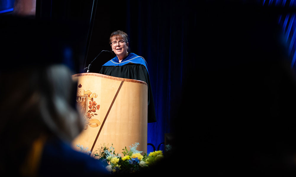 Person in academic regalia standing at a podium with an emblem, giving a speech at a graduation ceremony. Flowers are arranged in front of the podium.