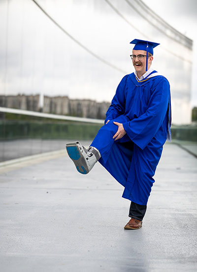 Person in a blue graduation gown and cap standing on one leg, with the other leg in a medical boot. The background shows an outdoor setting with modern architecture and blurred buildings.