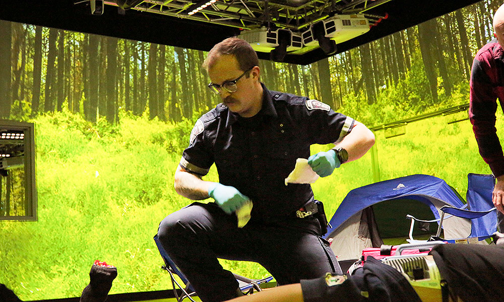 a male paramedic student in a training theatre that has been made to look like a campground. the student is kneeling over a patient