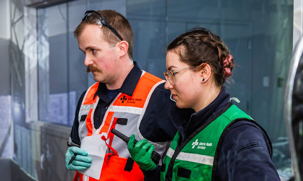 a man and a woman in safety vests stand side by side looking at something out of frame. the man holds a pen and piece of paper